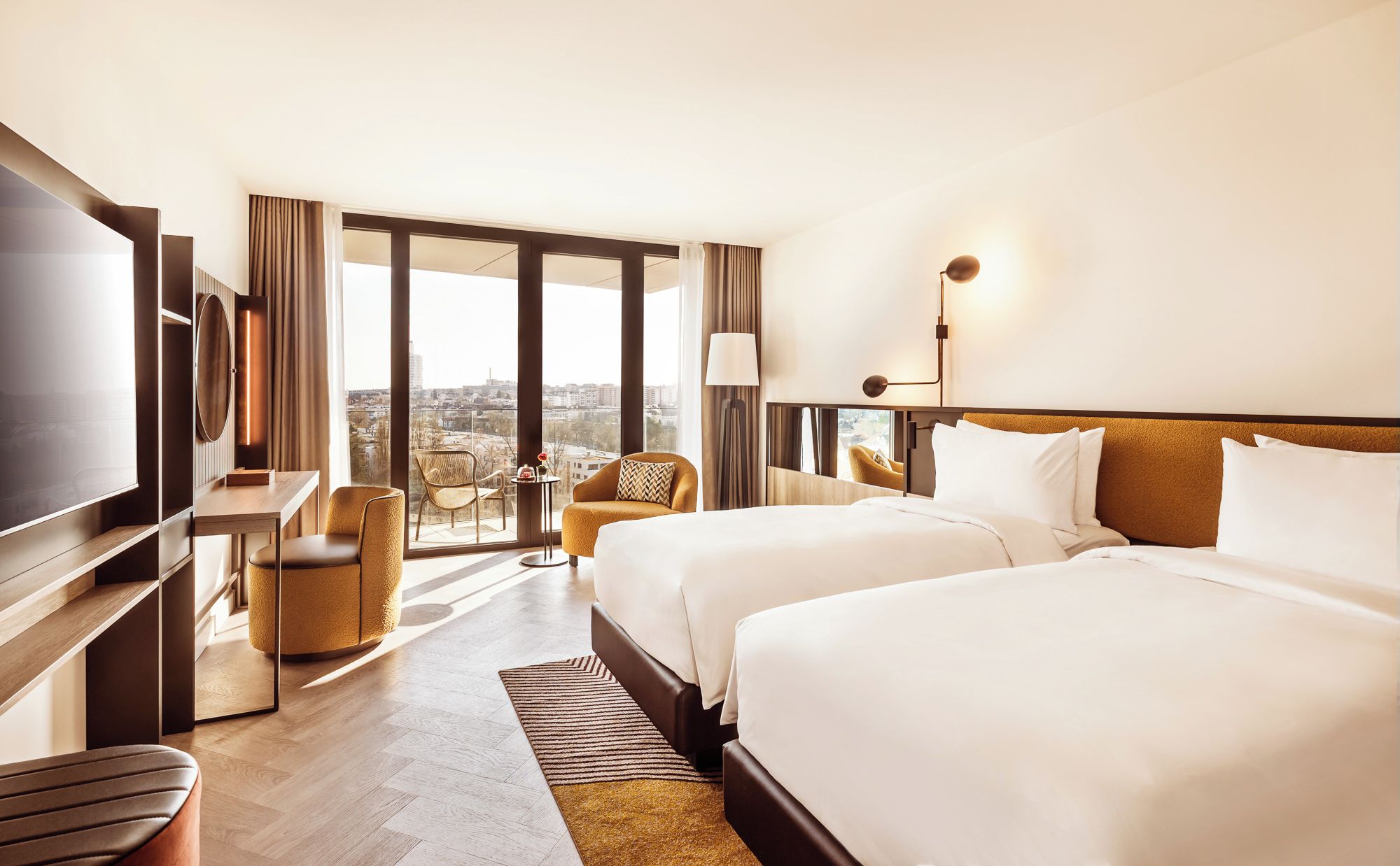 A hotel room with two beds, a desk, a chair, and a large window offering a city view.