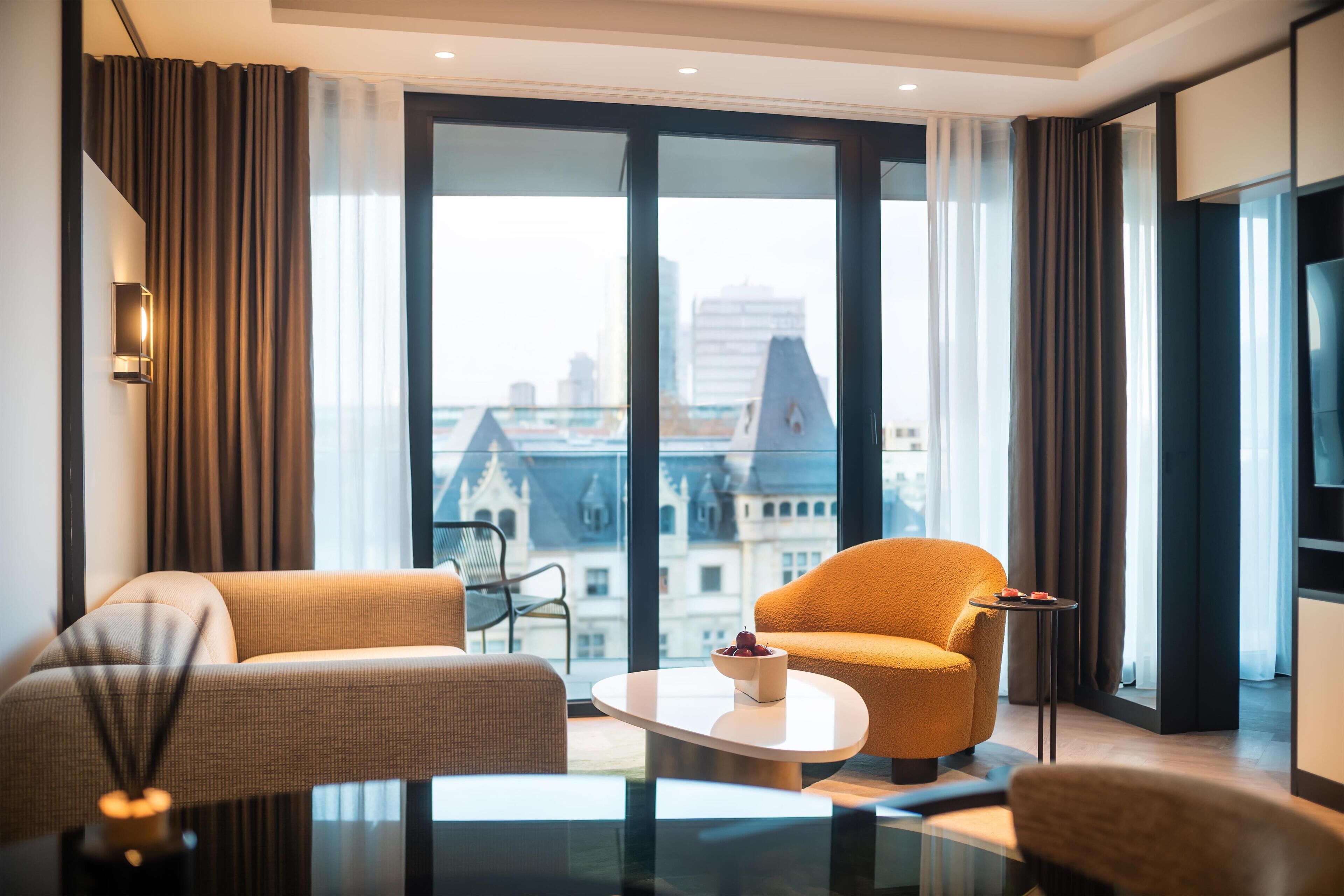 A modern hotel room with a large window offering a city view, a sofa, a chair, and a small table with a bowl of fruit.