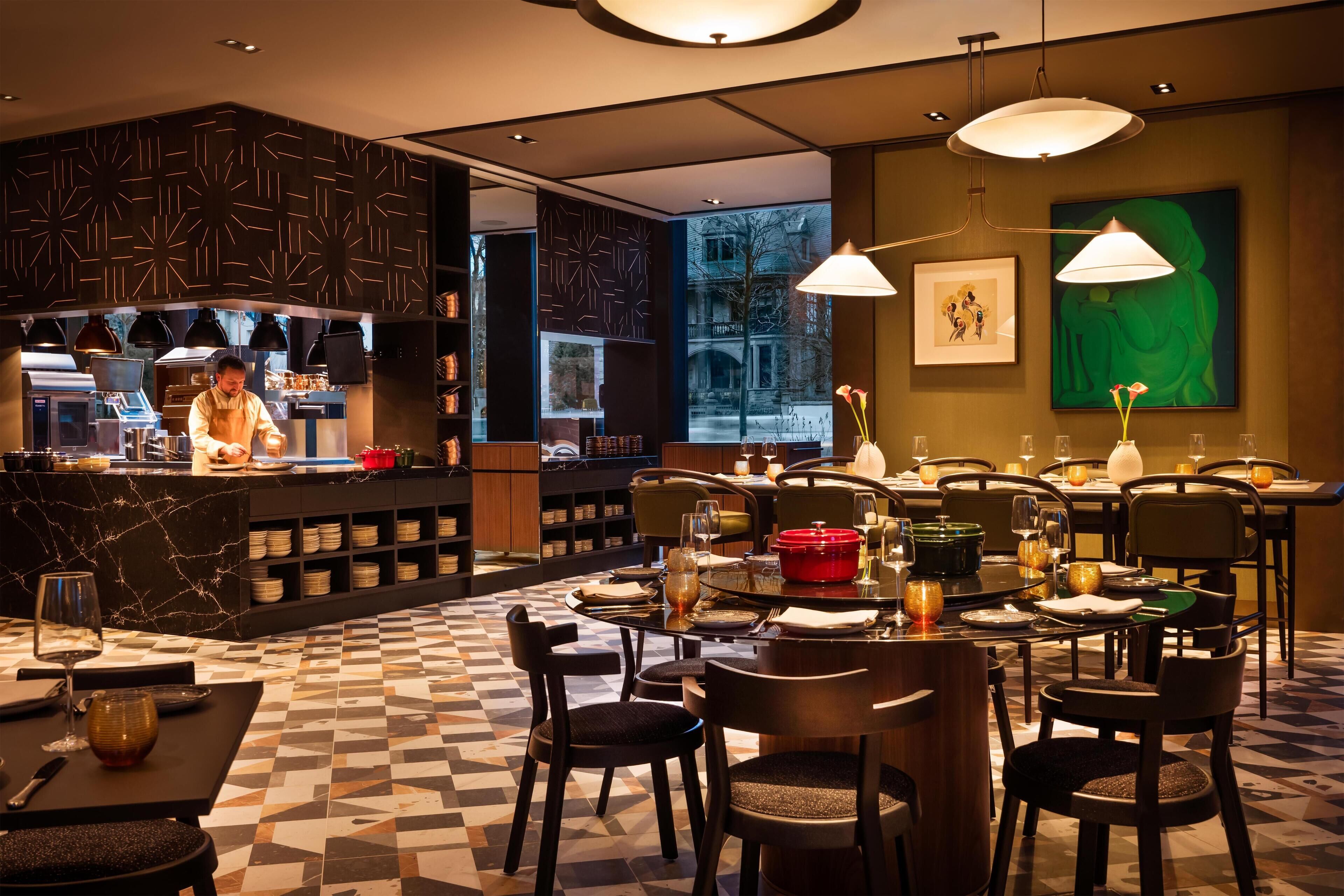 A restaurant with a checkered floor, tables set with wine glasses, plates, and a red pot. A chef is visible in the background.