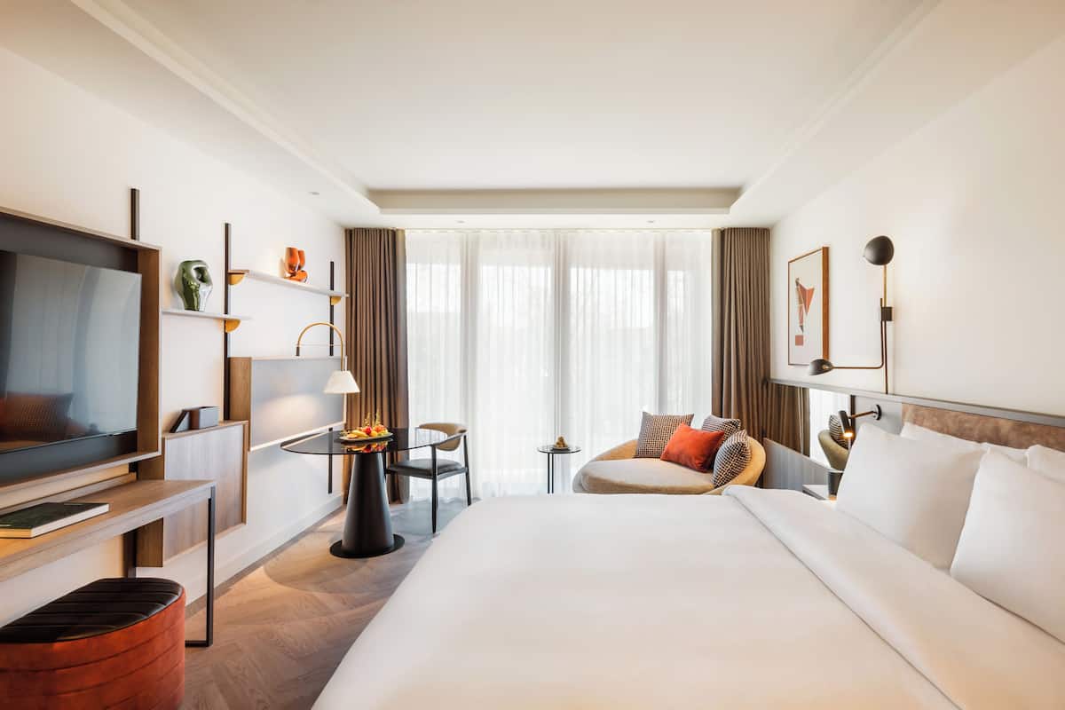 Una habitación de hotel con una cama grande, un televisor, un escritorio con lámpara, una mesita con fruta y una silla.