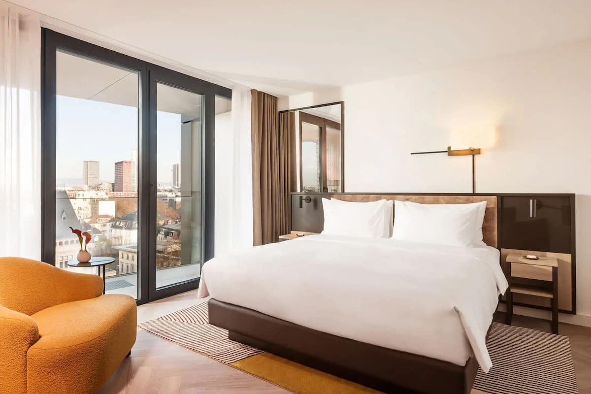 Una habitación de hotel con una cama grande, una silla, una mesita de noche y vistas de la ciudad a través de un ventanal amplio.