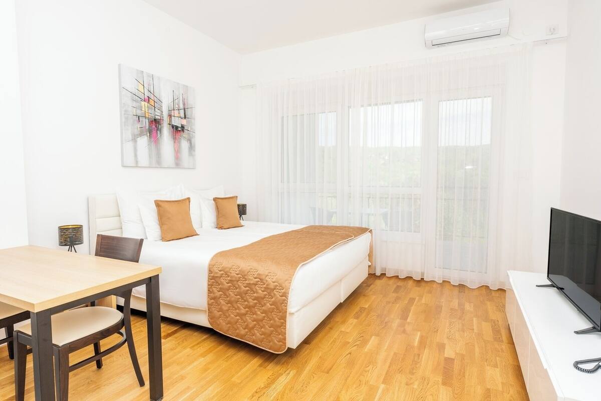 Une chambre d’hôtel avec un lit, un bureau, une chaise, une télévision et une fenêtre avec des rideaux légers.