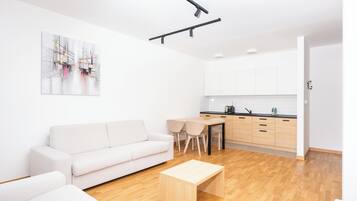 Una sala de estar moderna con un sofá, una mesa de centro y un comedor con sillas. Hay una cocina con armarios blancos y encimera negra.