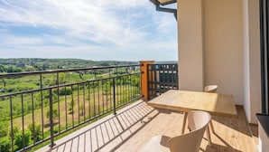 A balcony with a wooden table and chairs, a metal railing, and a view of a green landscape.