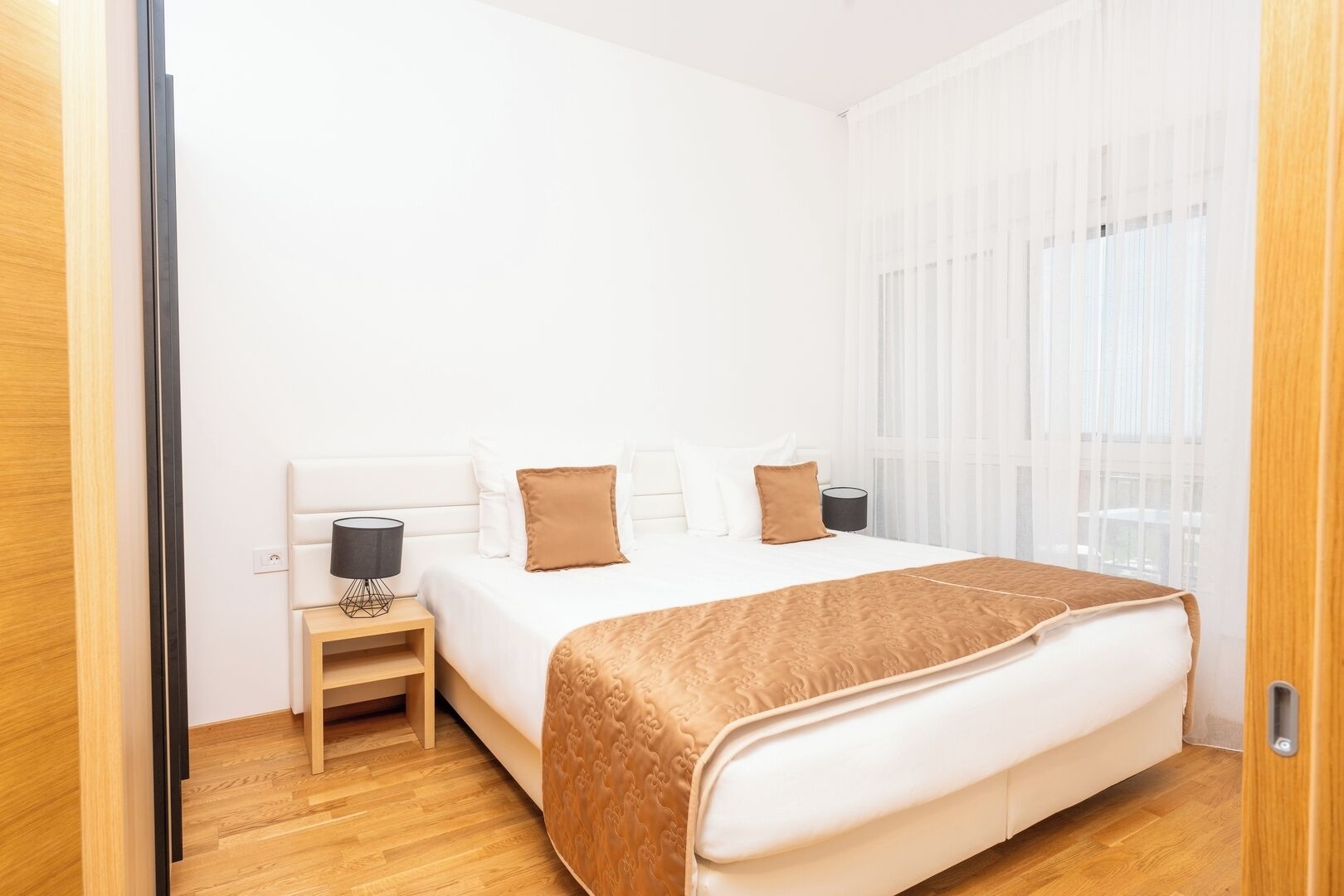 A bedroom with a large bed, a bedside table, and a window with sheer curtains.