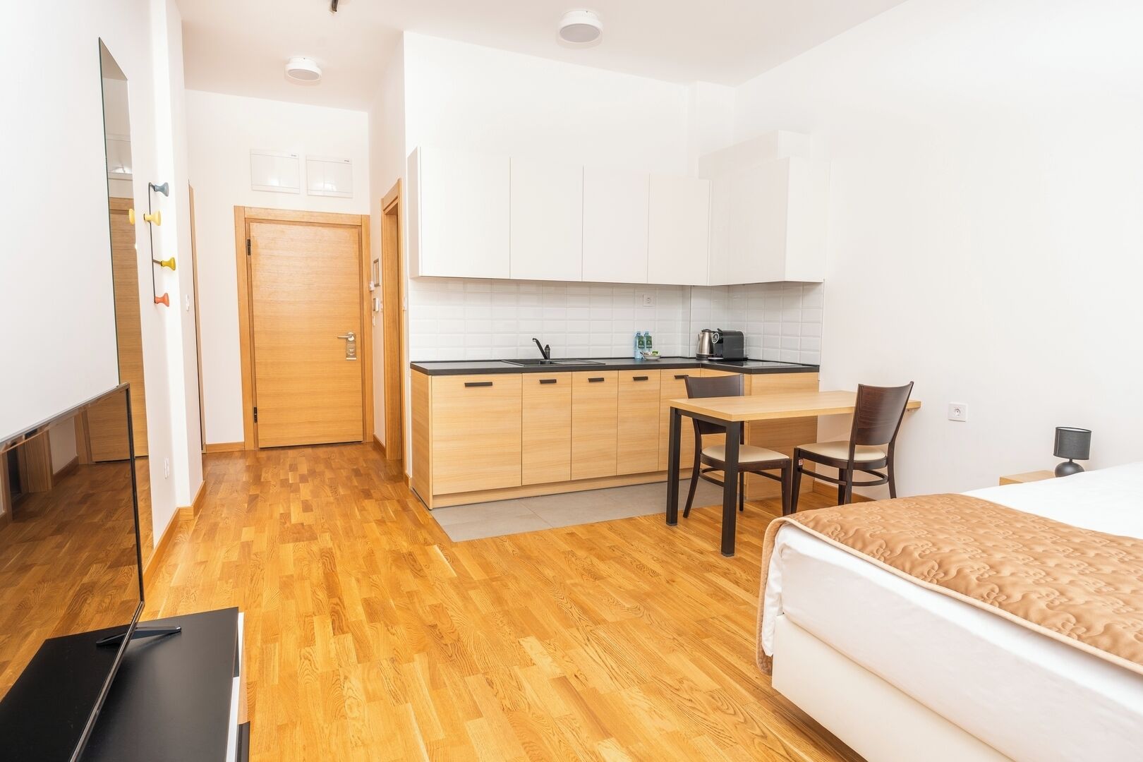 A compact hotel room with a wooden floor, a bed with a patterned cover, a small kitchenette with white cabinets, a dining table with two chairs, and a door leading to another room.