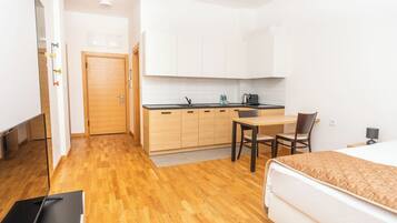 A compact hotel room with a wooden floor, a bed with a patterned cover, a small kitchenette with white cabinets, a dining table with two chairs, and a door leading to another room.