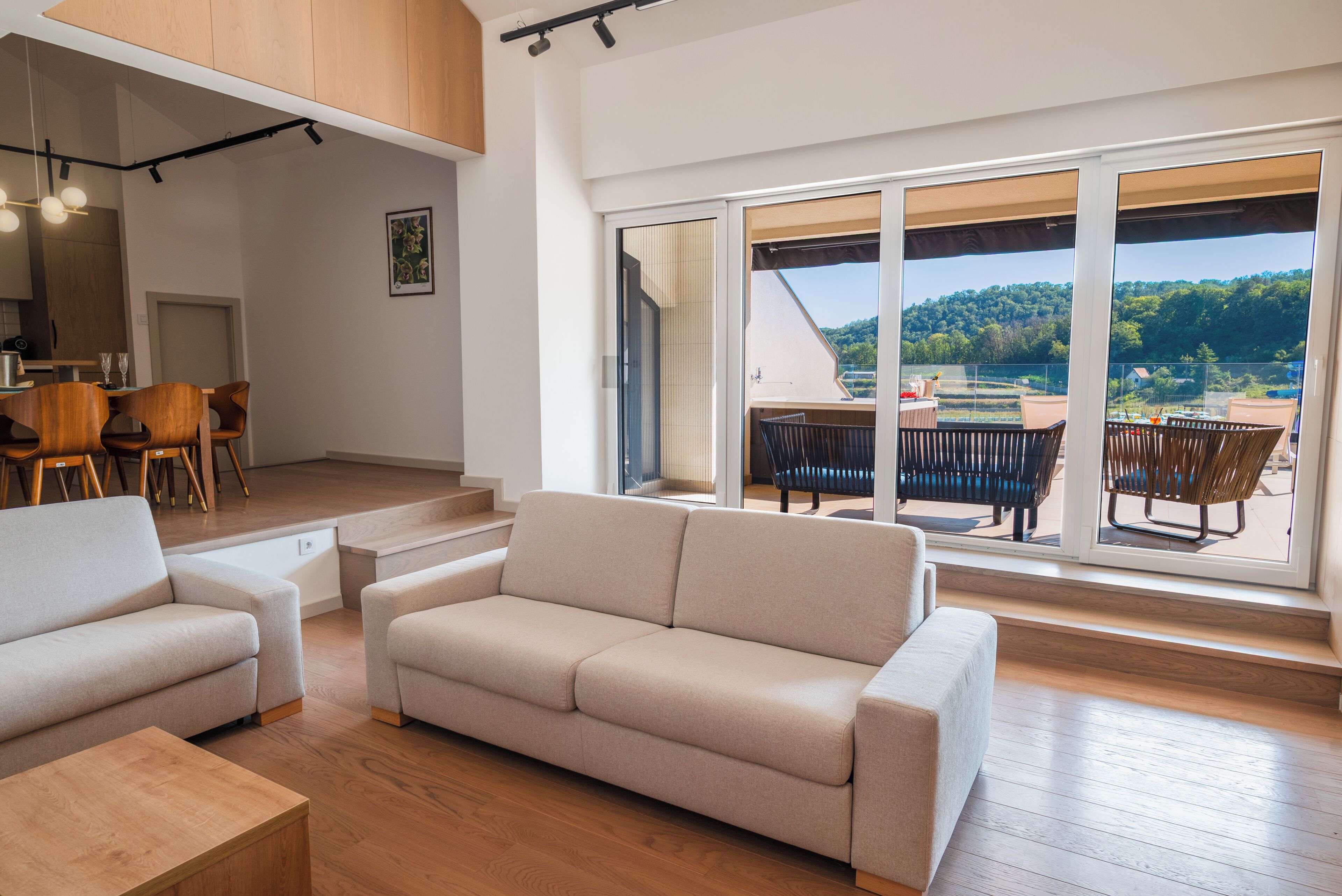 Un vaste espace de vie comprenant un canapé, une table à manger et des chaises, ainsi qu’une grande porte-fenêtre coulissante donnant sur une terrasse extérieure avec vue sur une colline boisée.