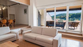 Un vaste espace de vie comprenant un canapé, une table à manger et des chaises, ainsi qu’une grande porte-fenêtre coulissante donnant sur une terrasse extérieure avec vue sur une colline boisée.