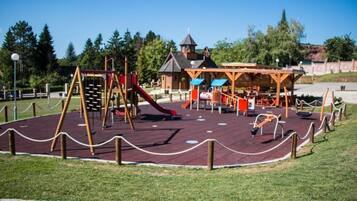 A playground with various play equipment, a covered seating area, and a building with a tower in the background.