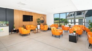 A modern reception area with orange seating, a wooden accent wall, a mounted TV, and large windows offering a view of greenery.