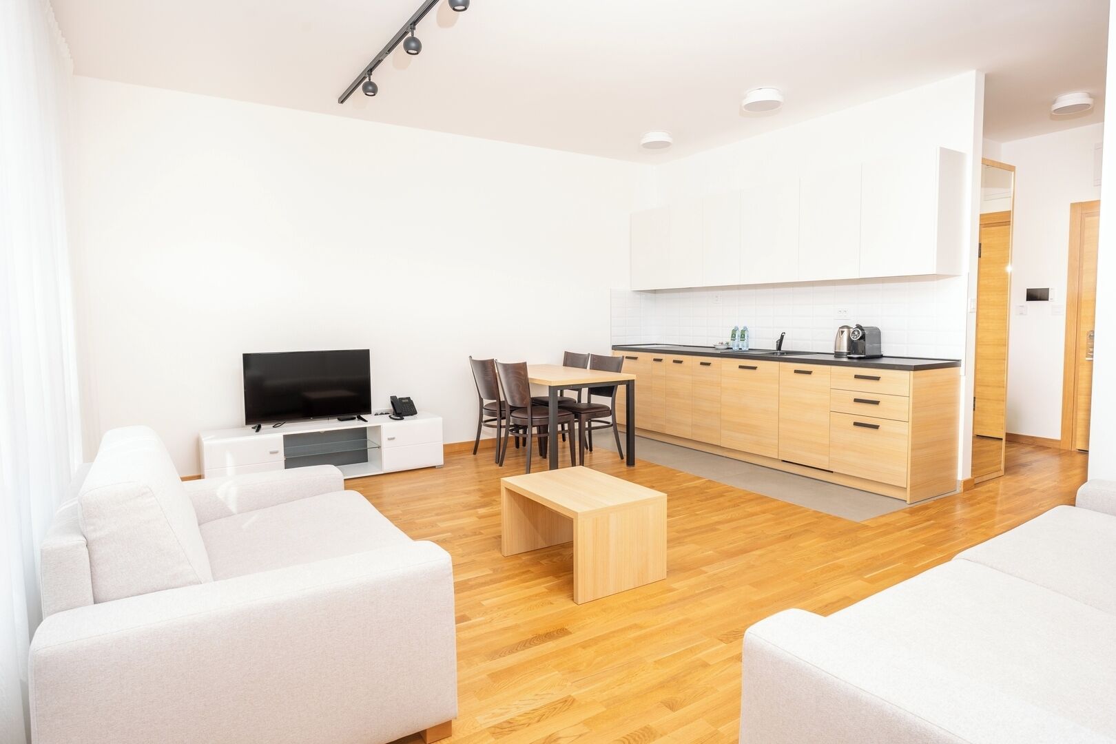 Una sala moderna con televisor de pantalla plana, sofá y mesa de centro. Hay una zona de comedor con sillas y una cocina con armarios y encimera.