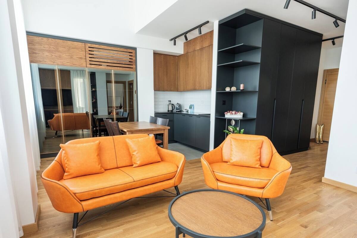 Un salon moderne avec un canapé et un fauteuil, un coin repas avec une table et des chaises, et une cuisine équipée d’armoires et d’un plan de travail.