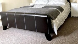 Cama bem arrumada com cabeceira preta e roupa de cama cinza, duas luminárias de mesa e piso coberto por carpete.