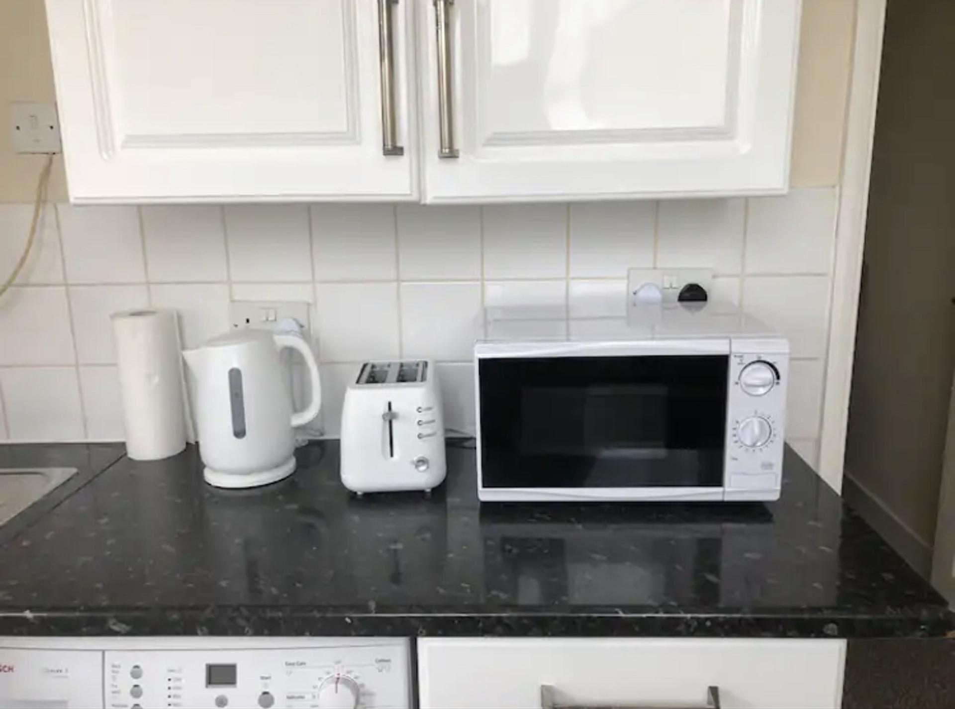 Eine Küche mit weißen Schränken, einem Toaster, einem Wasserkocher und einer Mikrowelle auf einer schwarzen Arbeitsplatte.