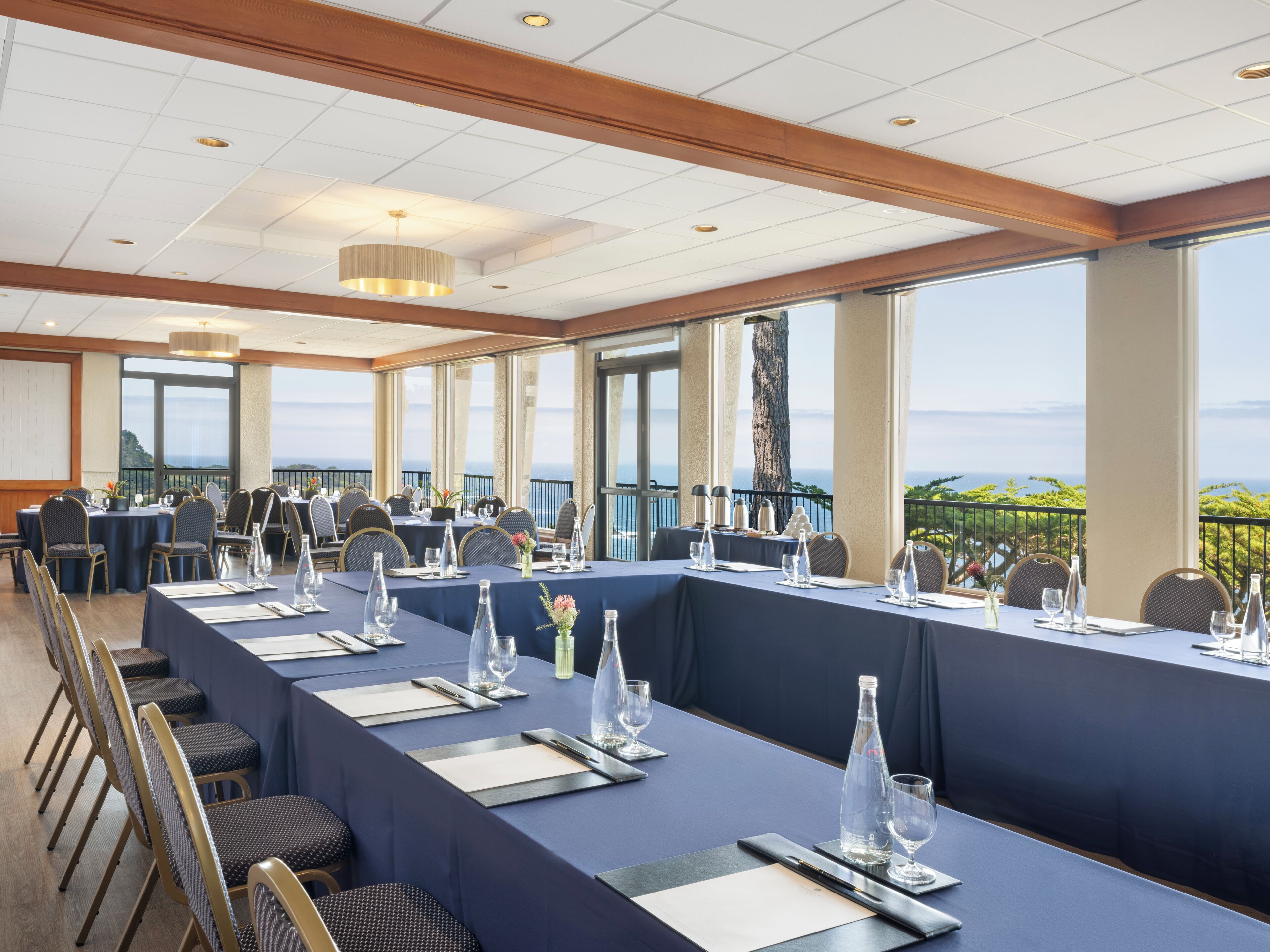 A conference room with a long table set for a meeting, large windows with a view of the sea, and a balcony with a railing.