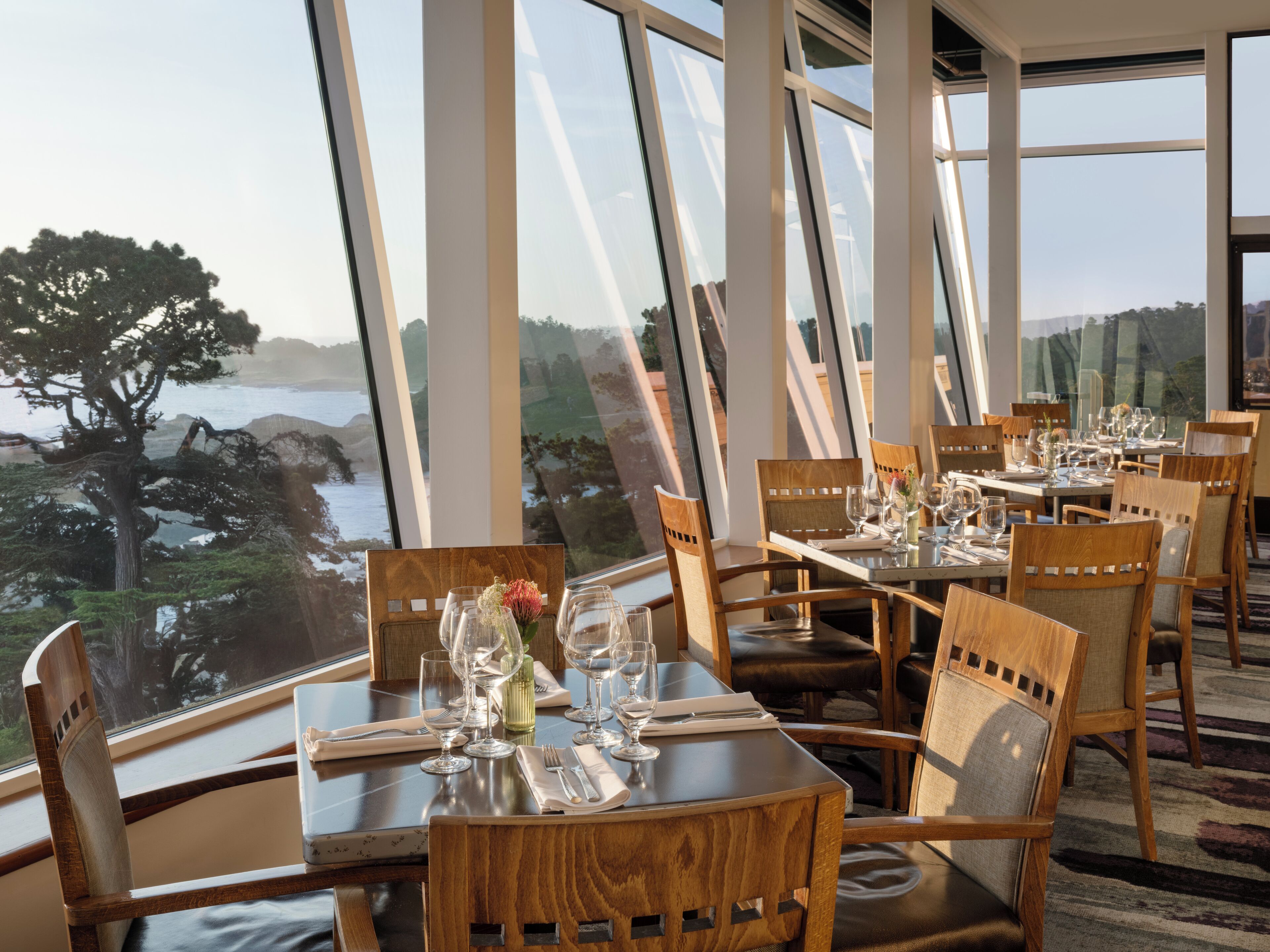 Comedor con mesas dispuestas para una comida, amplias ventanas que ofrecen vistas al mar y árboles, y sillas de madera.