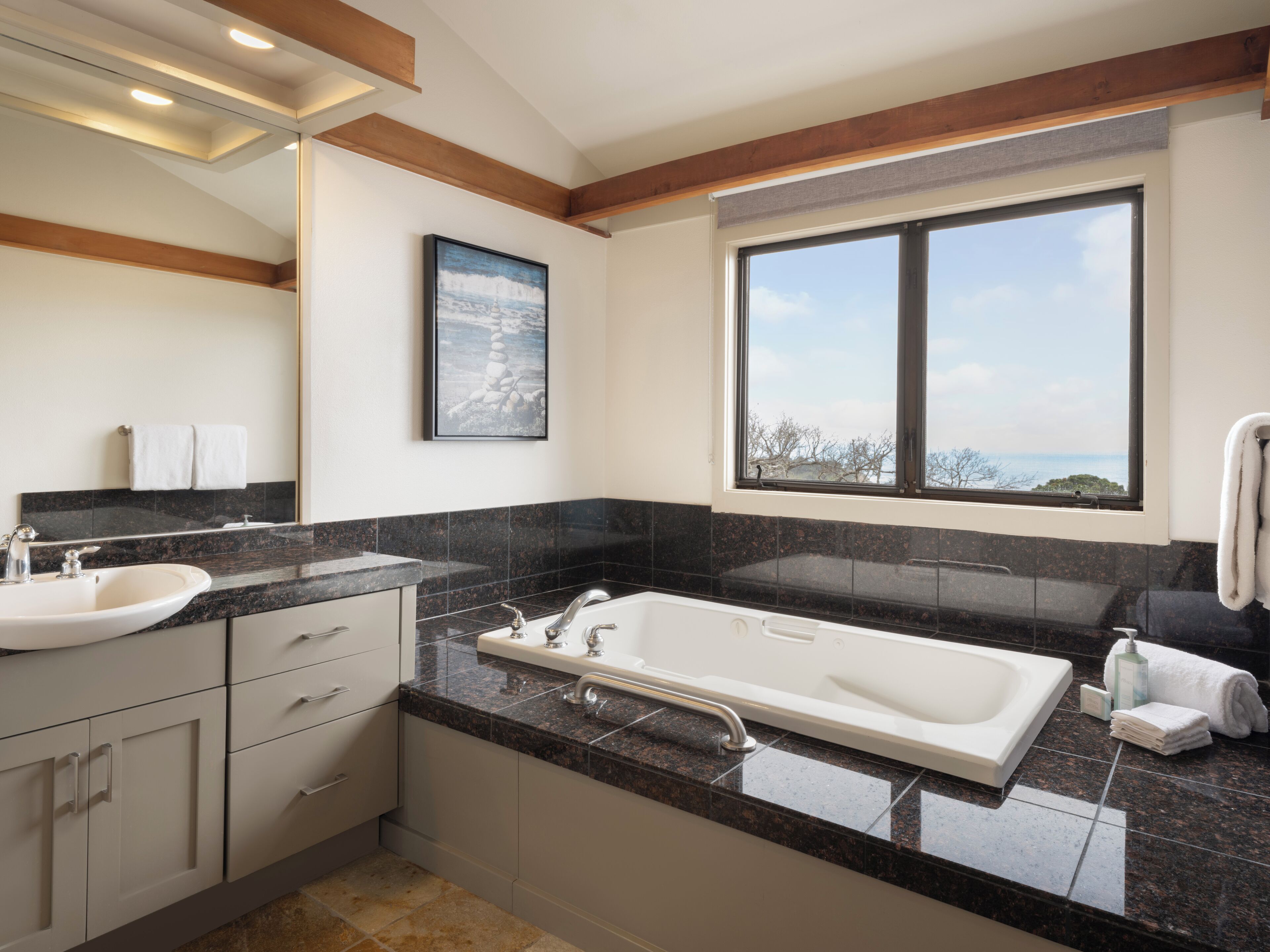 Un baño moderno con una bañera grande, un lavabo y un tocador. Hay una ventana con vistas a árboles y un cielo despejado.
