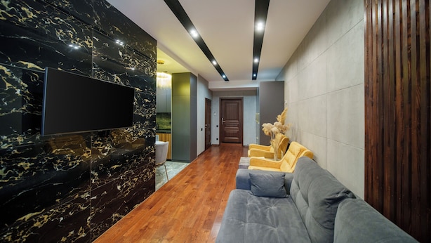 A modern living room with a flat-screen TV mounted on a marble wall, a wooden floor, a grey sofa, and a wooden panel on the right. - Arch Apart Hotel (Yerevan)