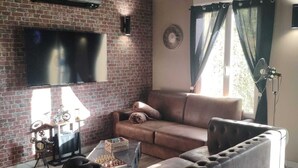 A living room with a brick wall, a flat-screen TV, a brown leather sofa, and a chessboard.