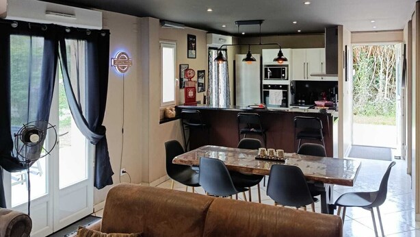 A modern living room with a brown sofa, dining table, and chairs, a kitchen area with cabinets and a refrigerator, and a large window with curtains.