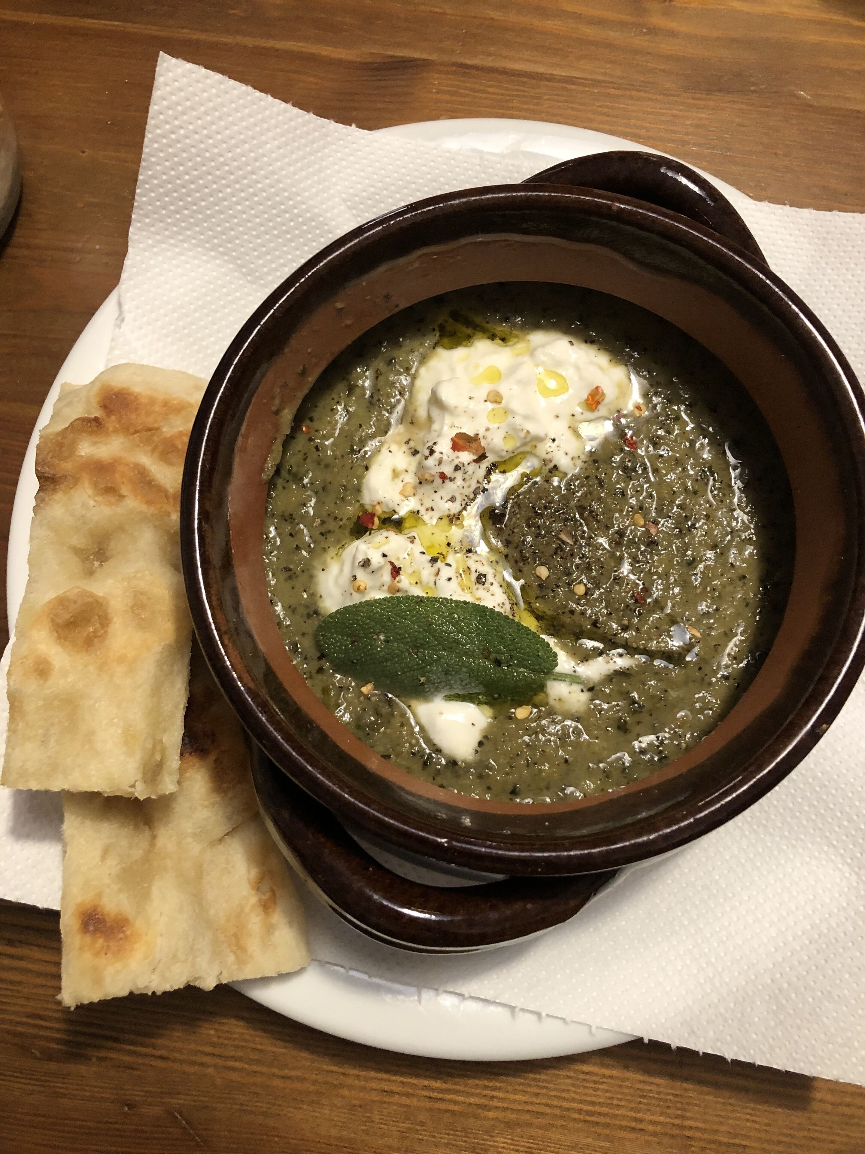 Un tazón de sopa con una cucharada de crema y una hoja verde encima, acompañado de un trozo de pan plano sobre un plato blanco.
