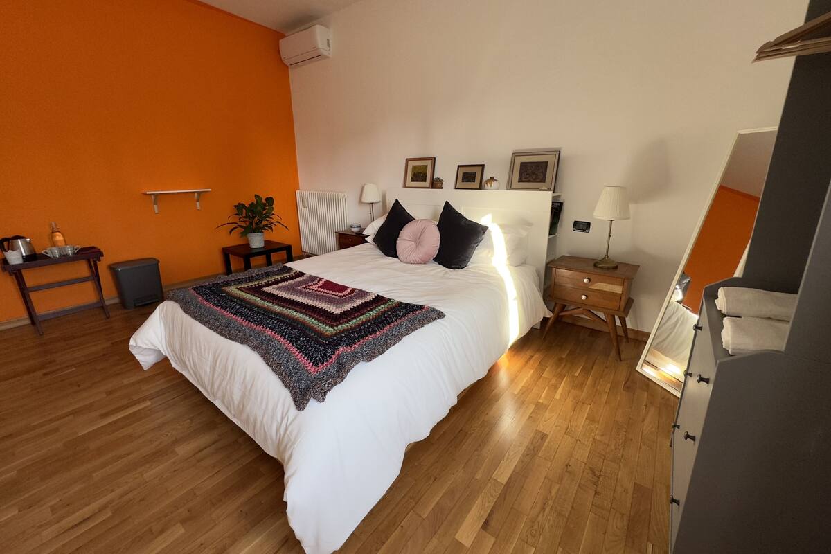 A modern bedroom with a large bed, a bedside table, a nightstand, a lamp, a wall-mounted air conditioner, and a potted plant.
