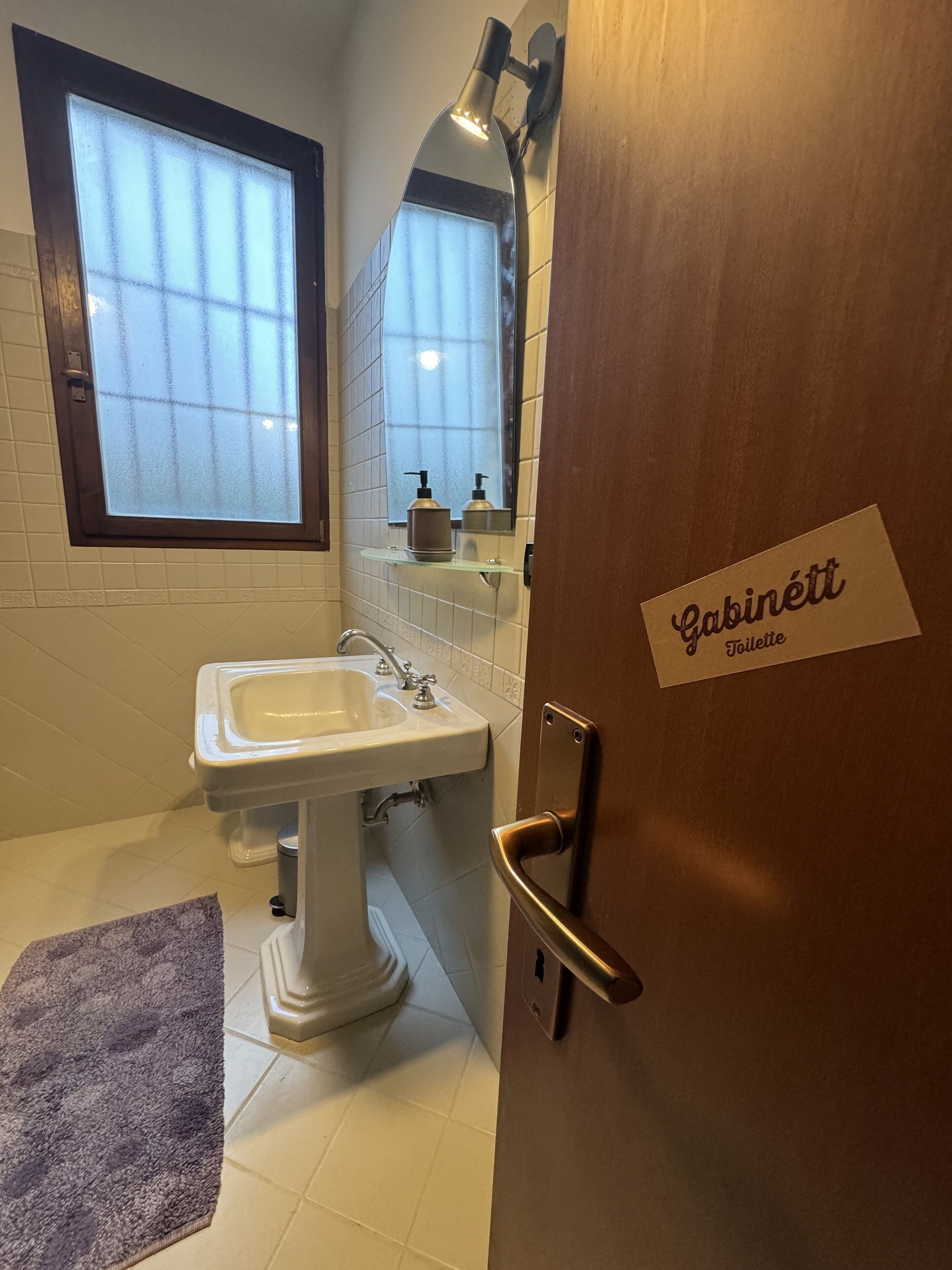 A bathroom with a white sink, a window, a mirror, and a door labeled "Cabinett Toilette".