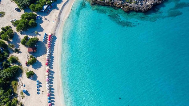 Eine Luftaufnahme eines Strandes mit klarem, türkisblauem Wasser, einer Reihe von Strandsonnenschirmen und einem Felsen.