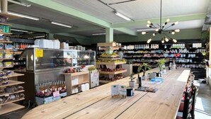 Ein geräumiger Laden mit einem zentralen Holztisch, Regalen voller verschiedener Produkte und einem Deckenventilator.