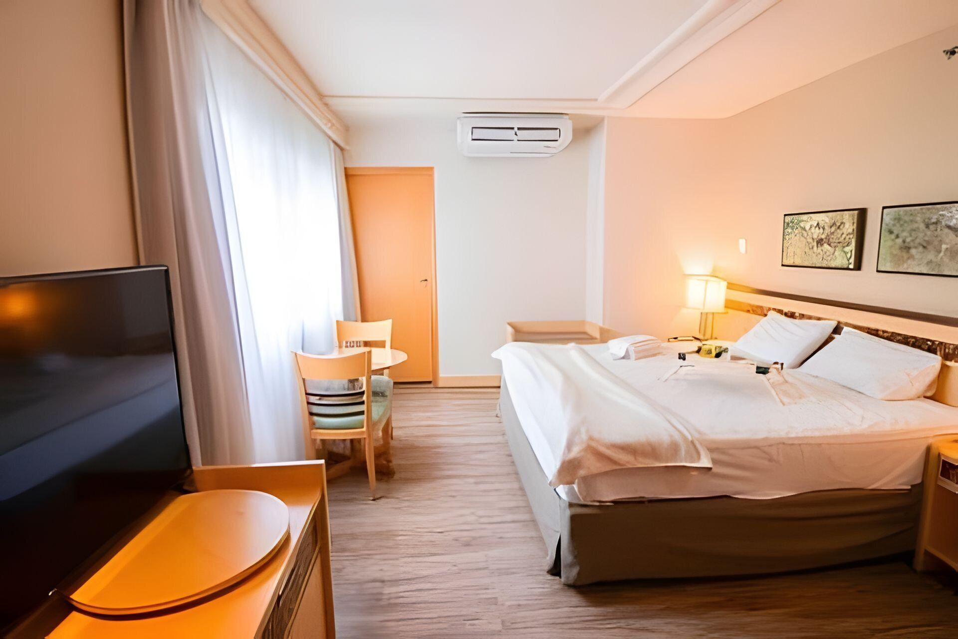 Habitación de hotel con cama, mesita de noche, lámpara, televisor, silla y puerta.