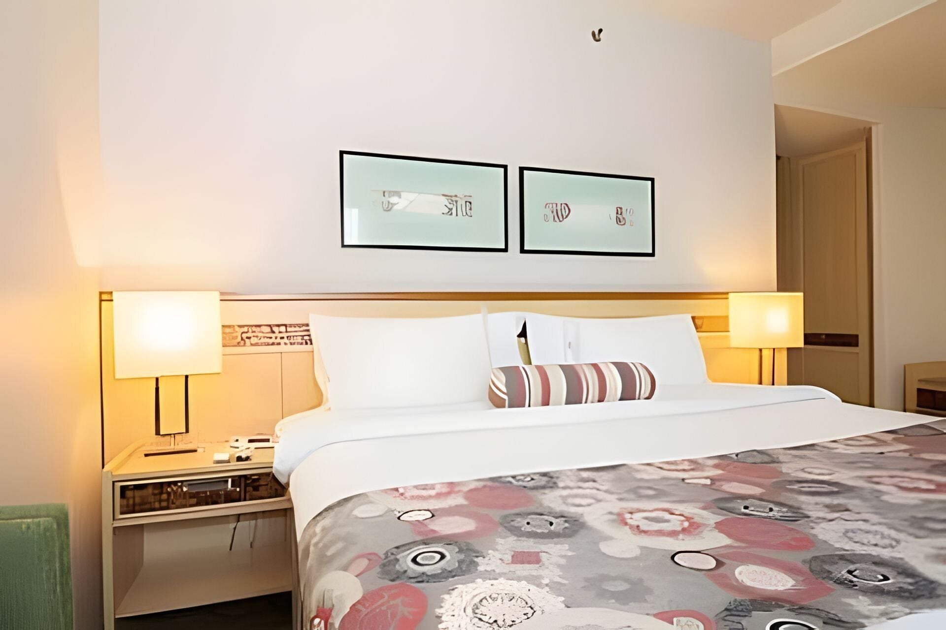 Una habitación de hotel con una cama, dos lámparas de noche, una mesita de noche y dos cuadros en la pared.