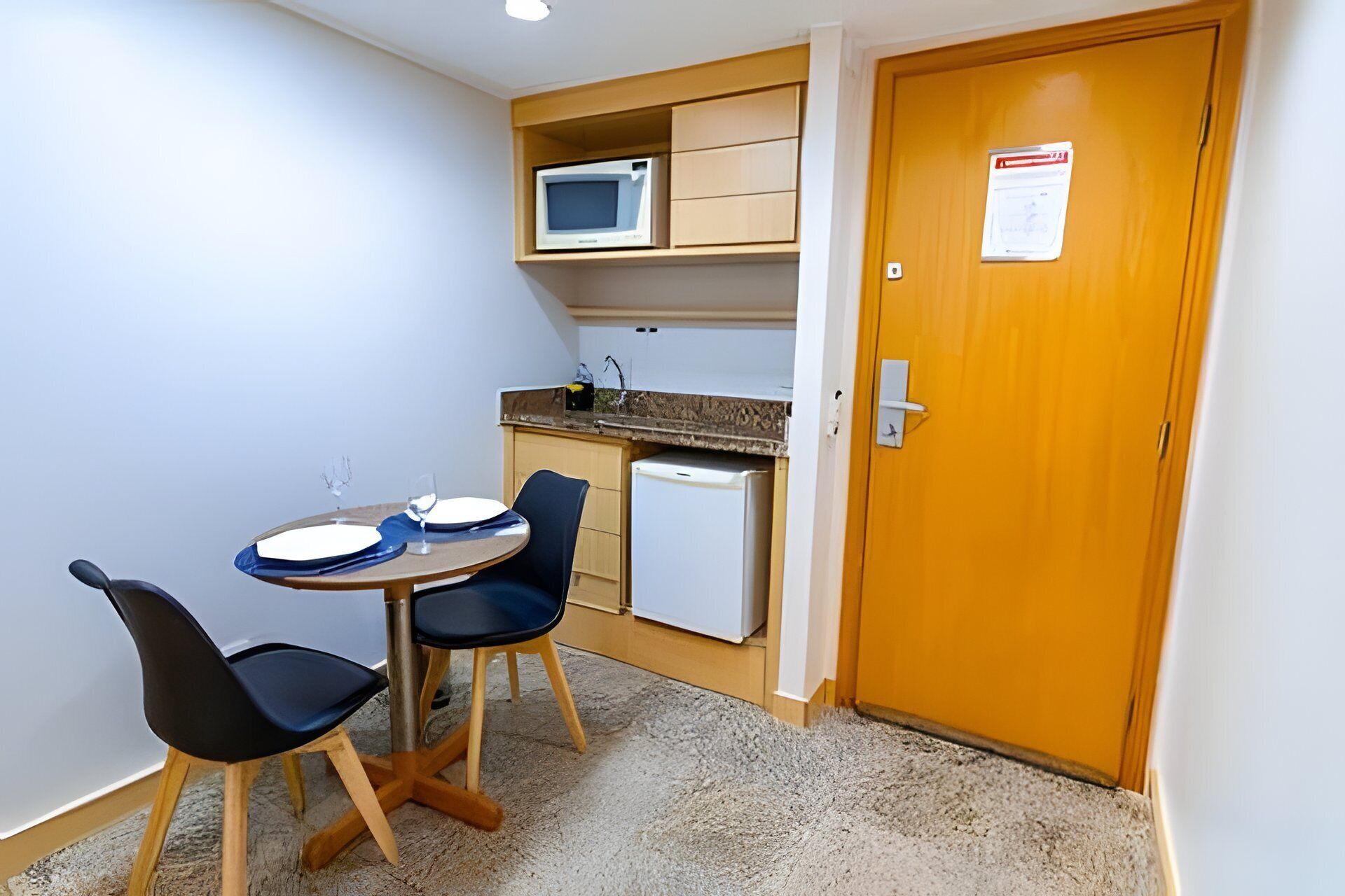 A small kitchenette with a microwave, a refrigerator, and a sink. There is a dining table with chairs and a small cabinet above it.
