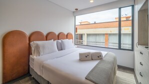 A modern hotel room with a large bed, white bedding, and a view of a building through the window.