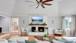 A spacious living room with a fireplace, a ceiling fan, and a large window.