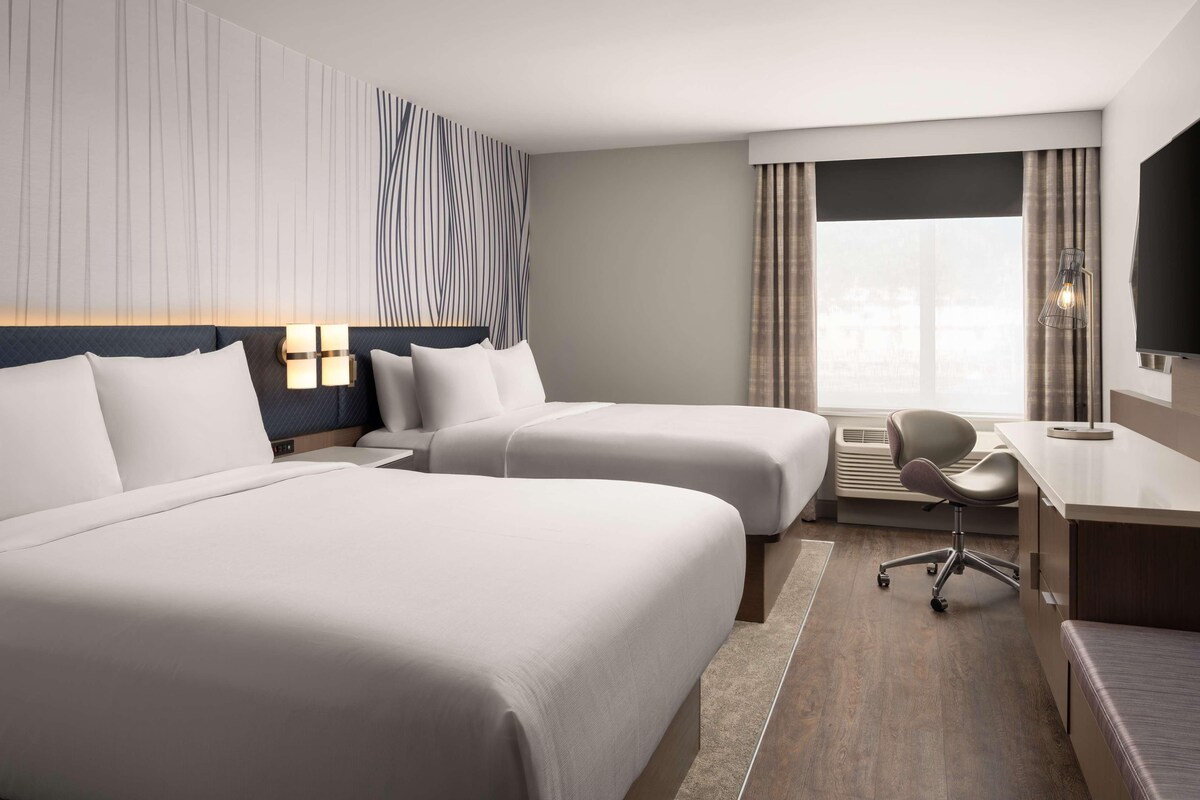 A hotel room with two beds, a desk, a chair, and a large window with curtains.