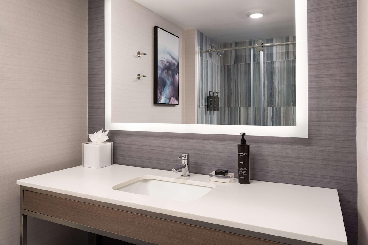 A modern bathroom with a large mirror, a white sink, a towel dispenser, and a shower with a glass door.