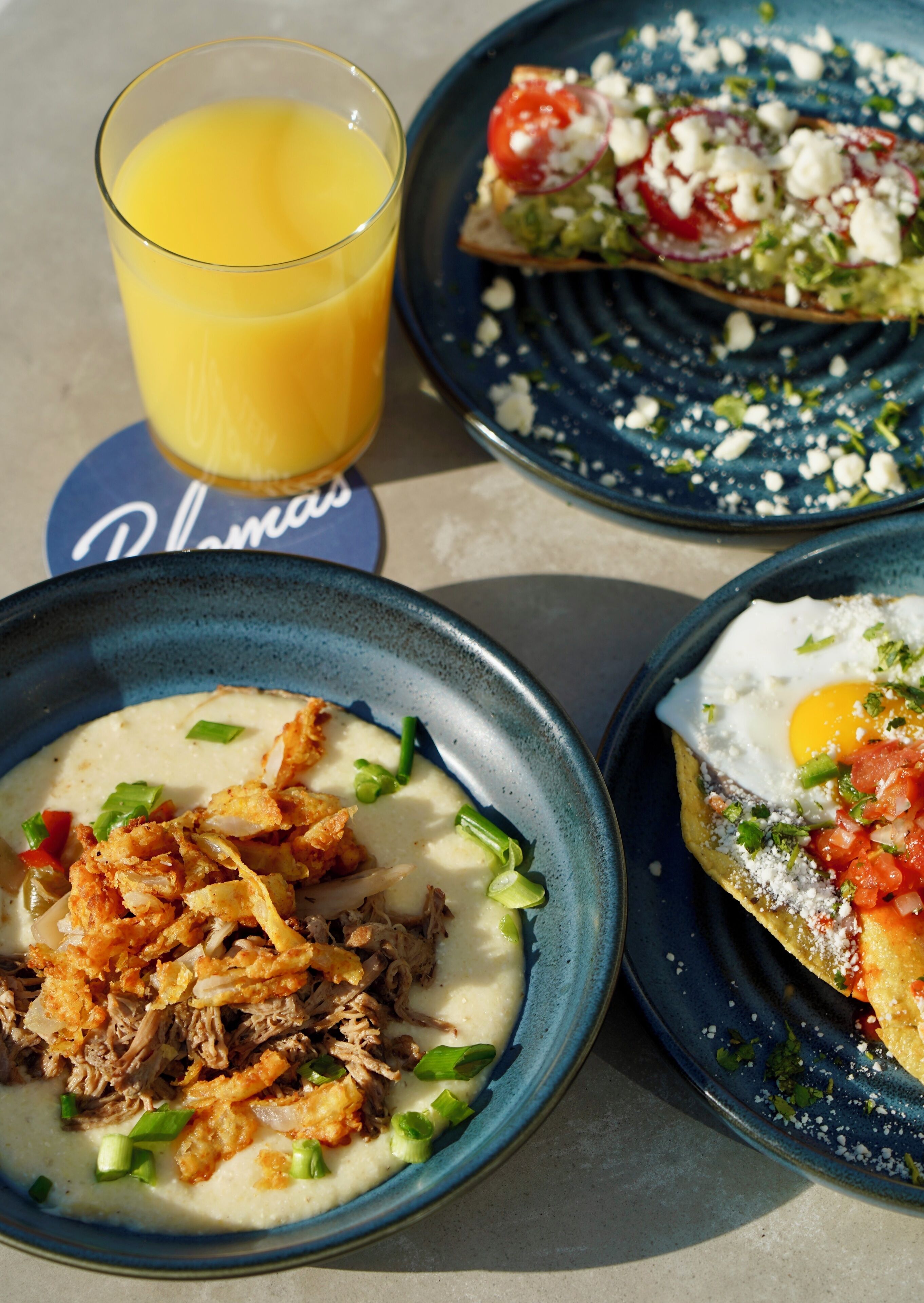Tres platos de comida, un vaso de zumo de naranja y un mantel individual con el nombre 'Plaza'.