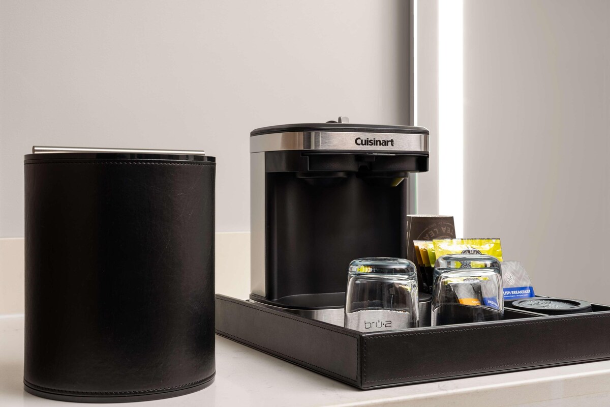 Un bolso de cuero negro, una cafetera Cuisinart y una bandeja con cristalería y cápsulas de café sobre una encimera.