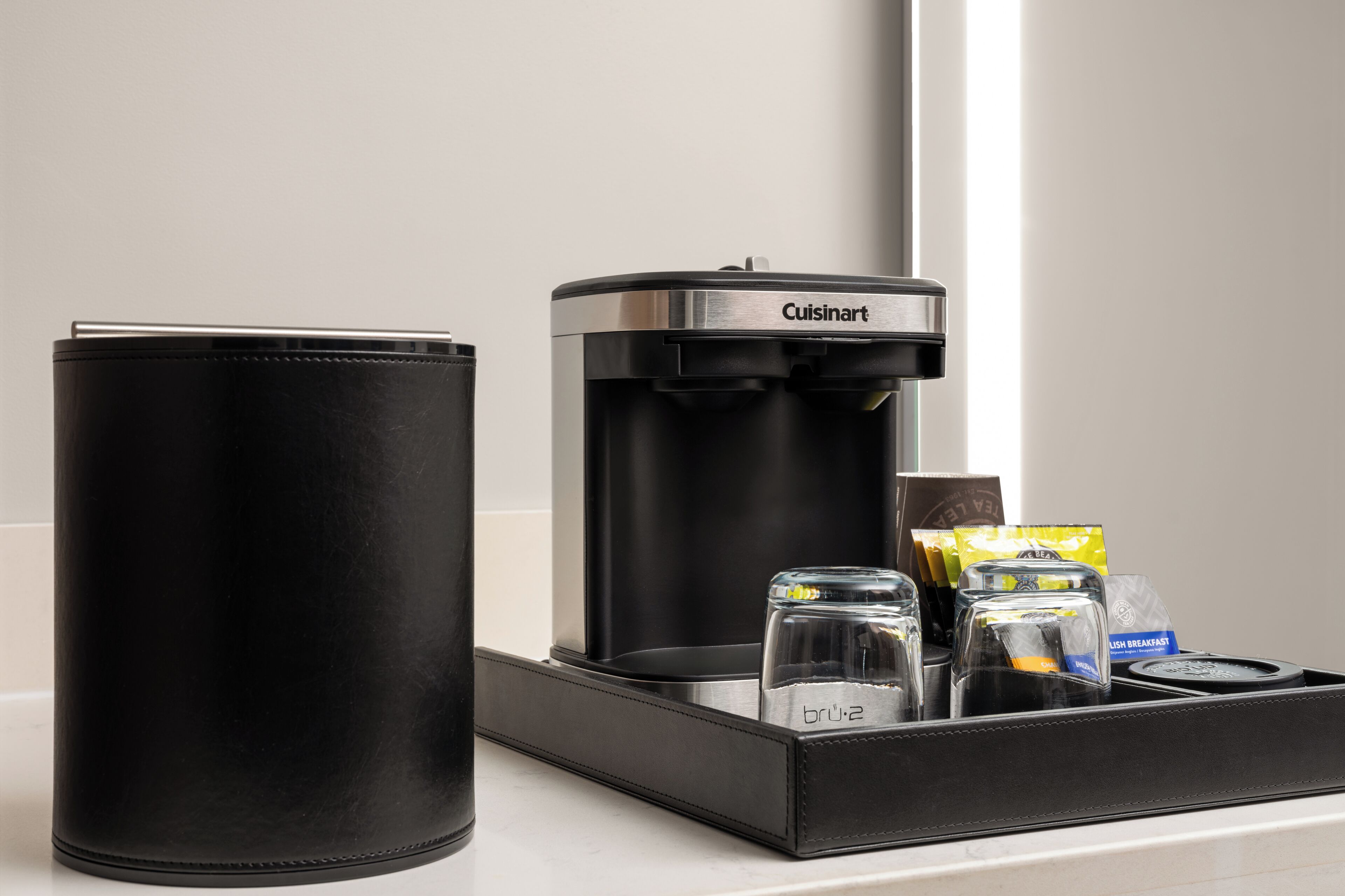 A black leather bag, a Cuisinart coffee maker, and a tray with glass containers and coffee pods on a countertop.