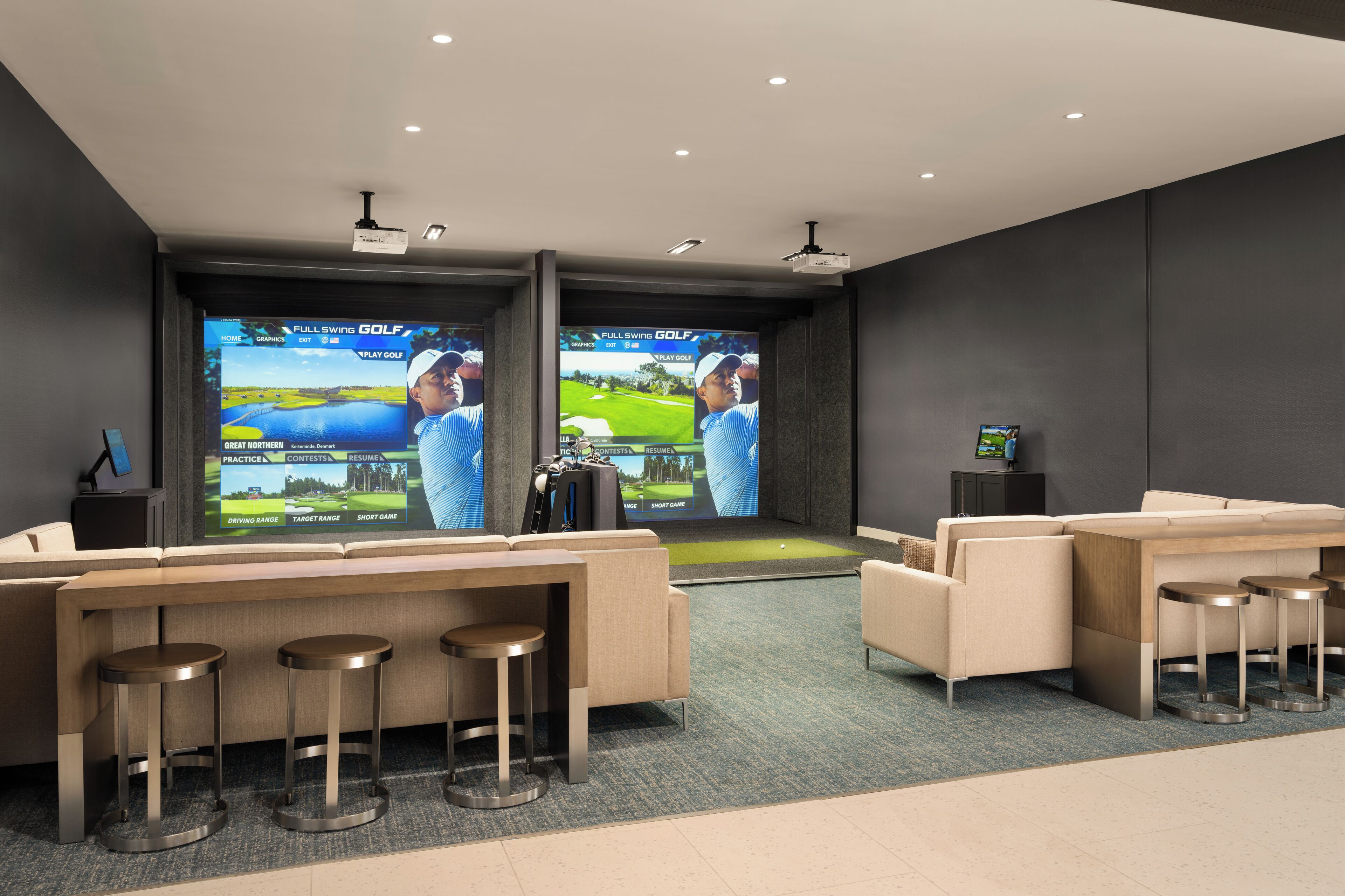 A modern lounge area with two large screens displaying golf content, comfortable seating, and a bar area.
