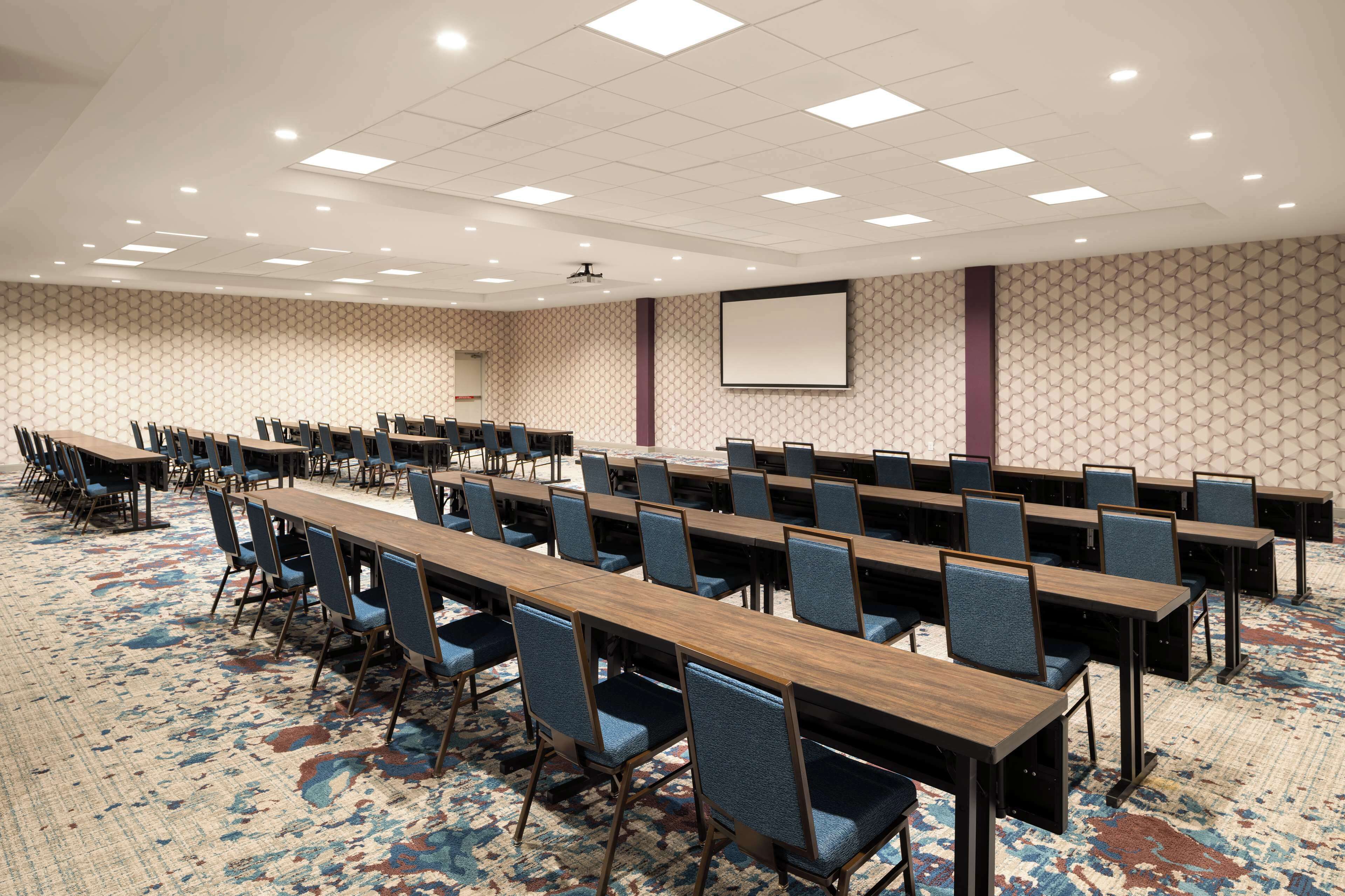 A conference room with long tables and chairs, a carpeted floor, and a projector screen on the wall.