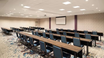 A conference room with long tables and chairs, a carpeted floor, and a projector screen on the wall.