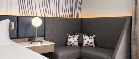 A modern hotel room with a grey sofa, a bedside table with a lamp, and a patterned wall.