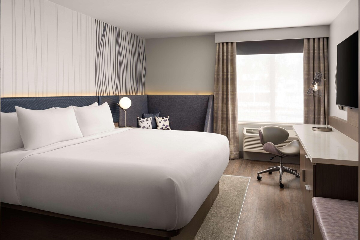 A hotel room with a large bed, a desk, a chair, a flat-screen TV, and a window with curtains.