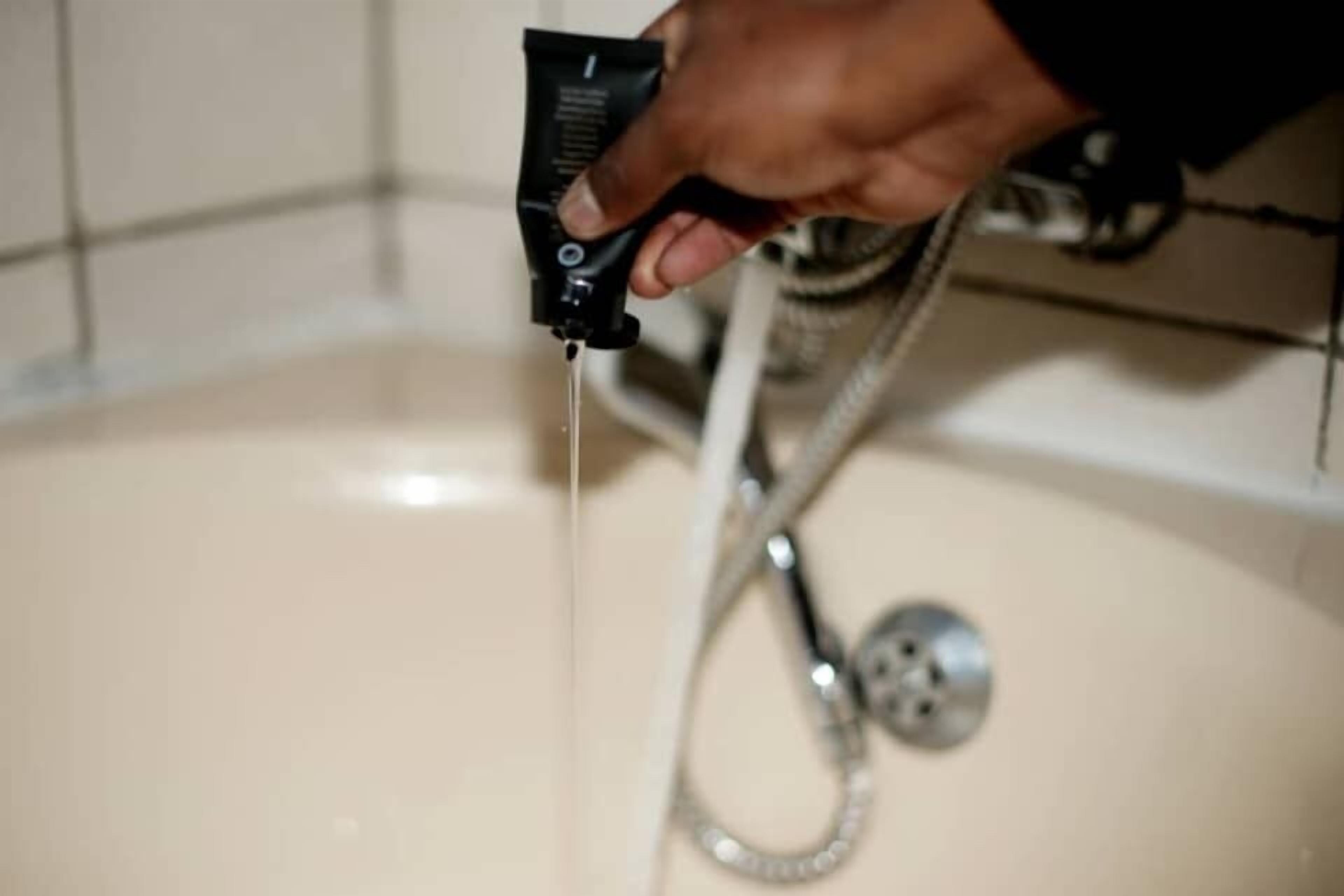 A hand holding a black device, possibly a razor, over a bathtub with a showerhead.