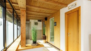 Un pasillo interior de madera con ventanas amplias, una puerta de madera y plantas en macetas.