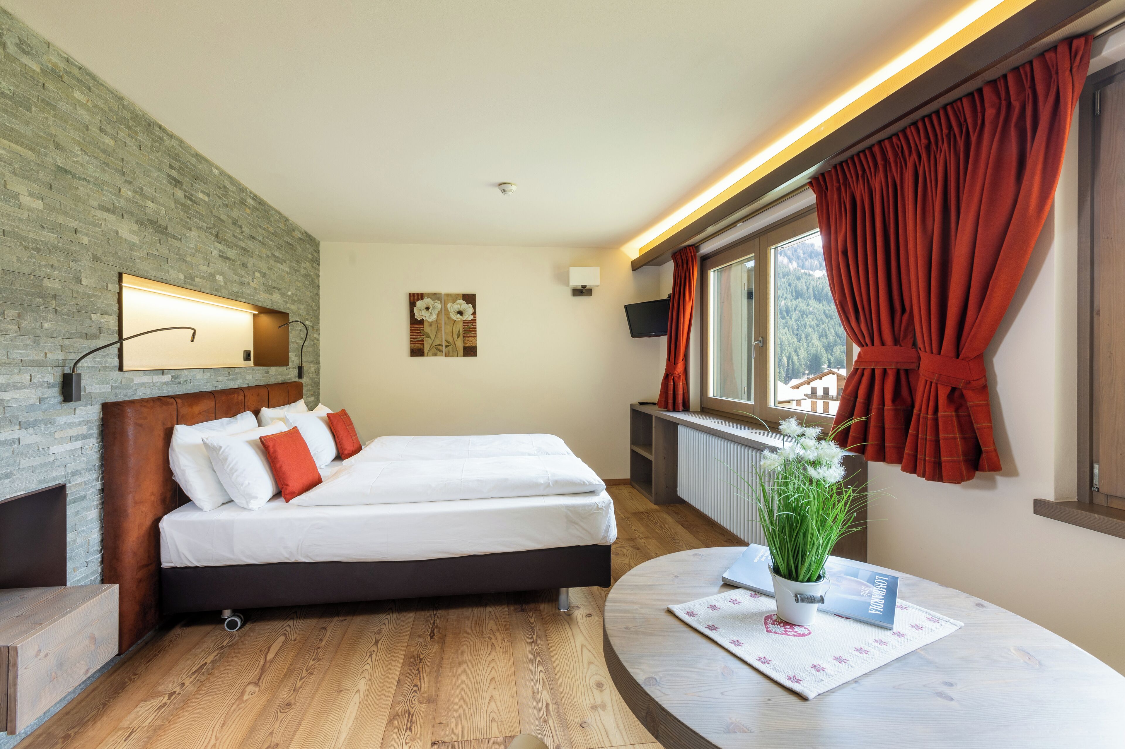 A modern bedroom with a wooden headboard, a bed with white and red bedding, a round table with a plant and a book, a brick-patterned wall, a mounted TV, and a window with red curtains.