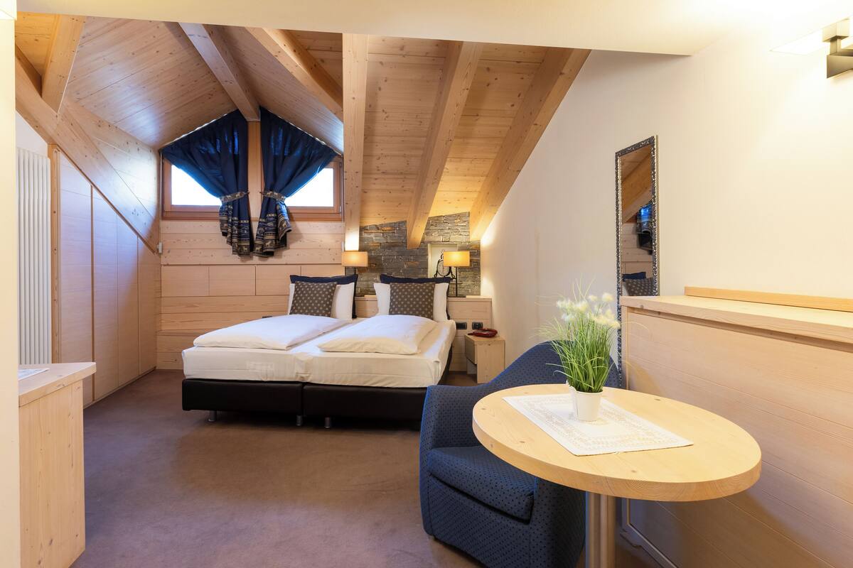 A bedroom with a wooden ceiling, a bed with white bedding, a blue chair, a small table with a plant, and a stone wall.