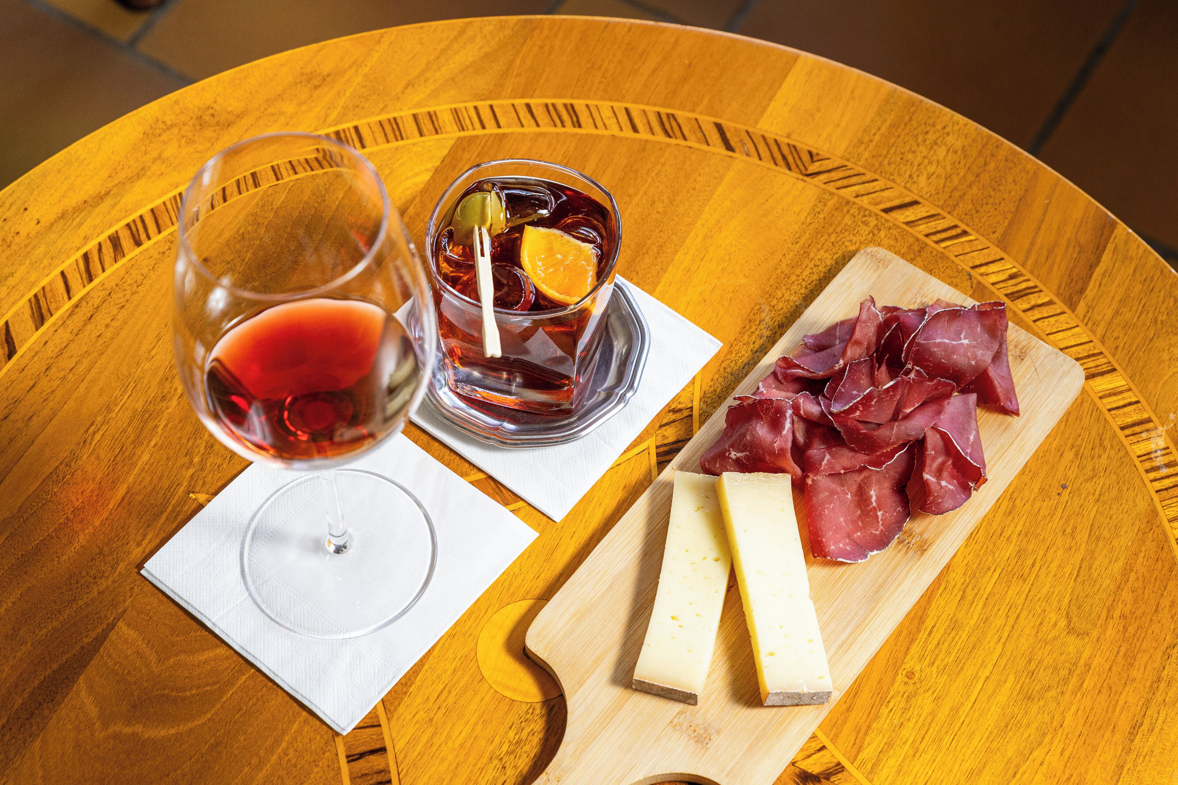 A round wooden table with a glass of red wine, a glass of iced tea with fruit, and a wooden board with cheese and cured meat.